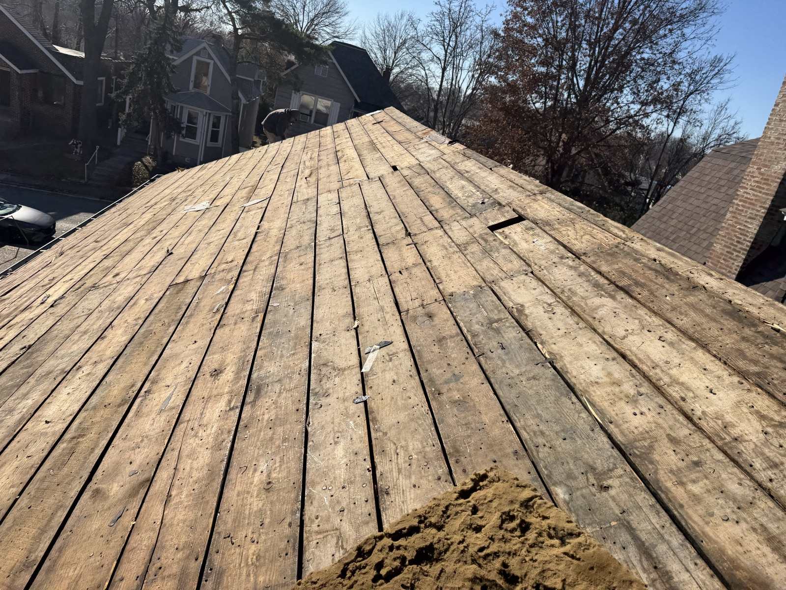 Old wooden plank decking exposed across a roof slope after a tear-off, showing nail holes, gaps between the boards and a pile of sawdust