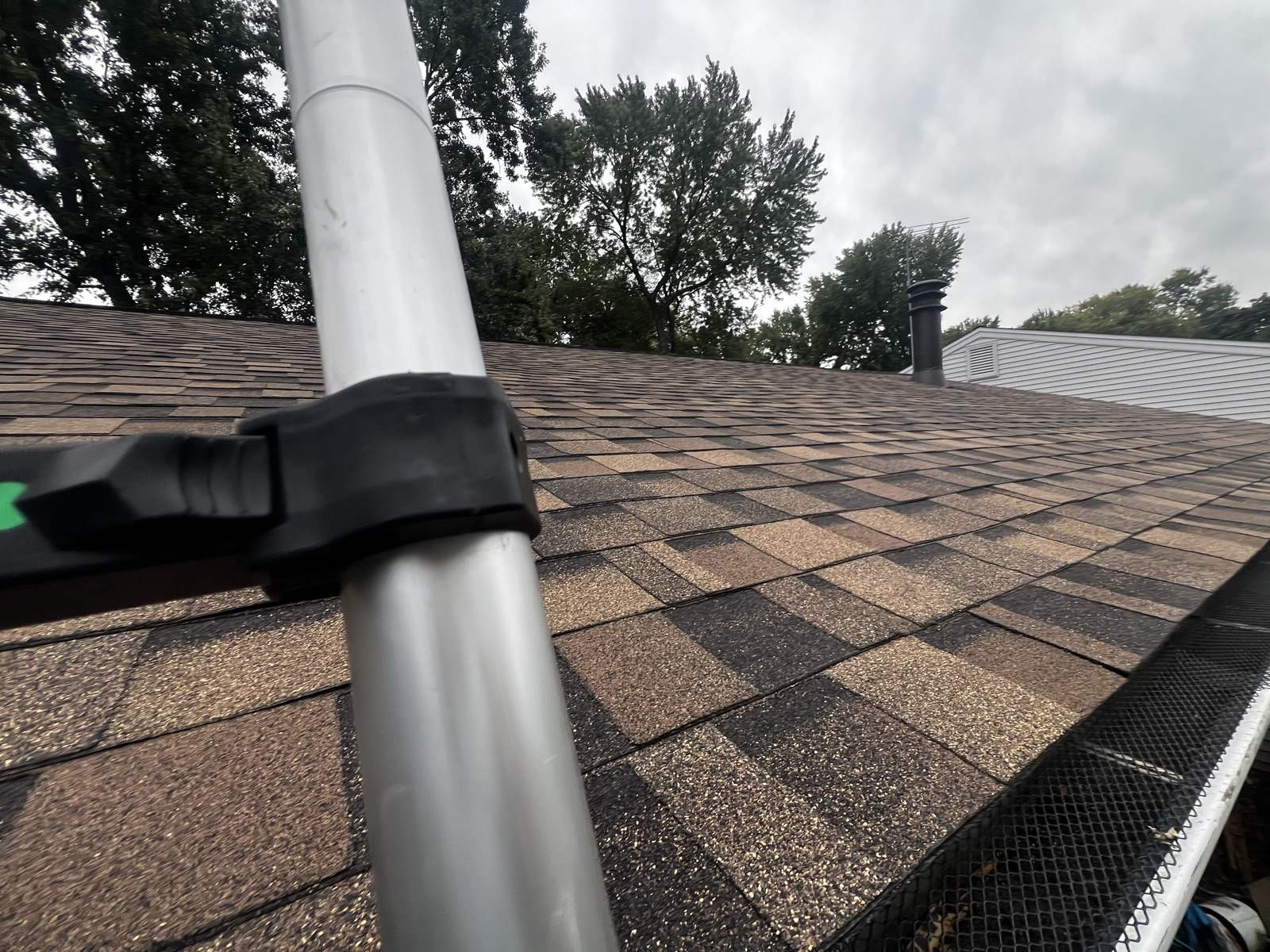 A weathered brown shingle roof under an overcast sky, with a black mesh gutter guard along the eave at the right and an aluminium extension pole crossing the frame
