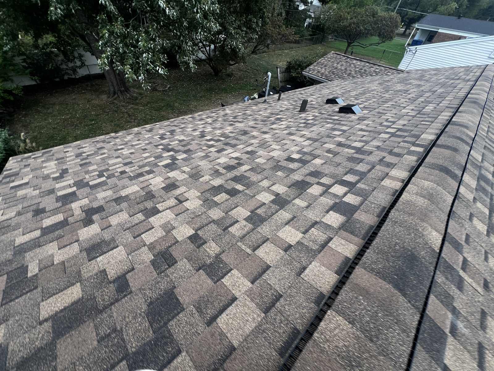 Broad expanse of a newly laid brown, tan and charcoal multi-tone architectural shingle roof with a capped ridge, a dark ridge vent, two box vents and a metal flue