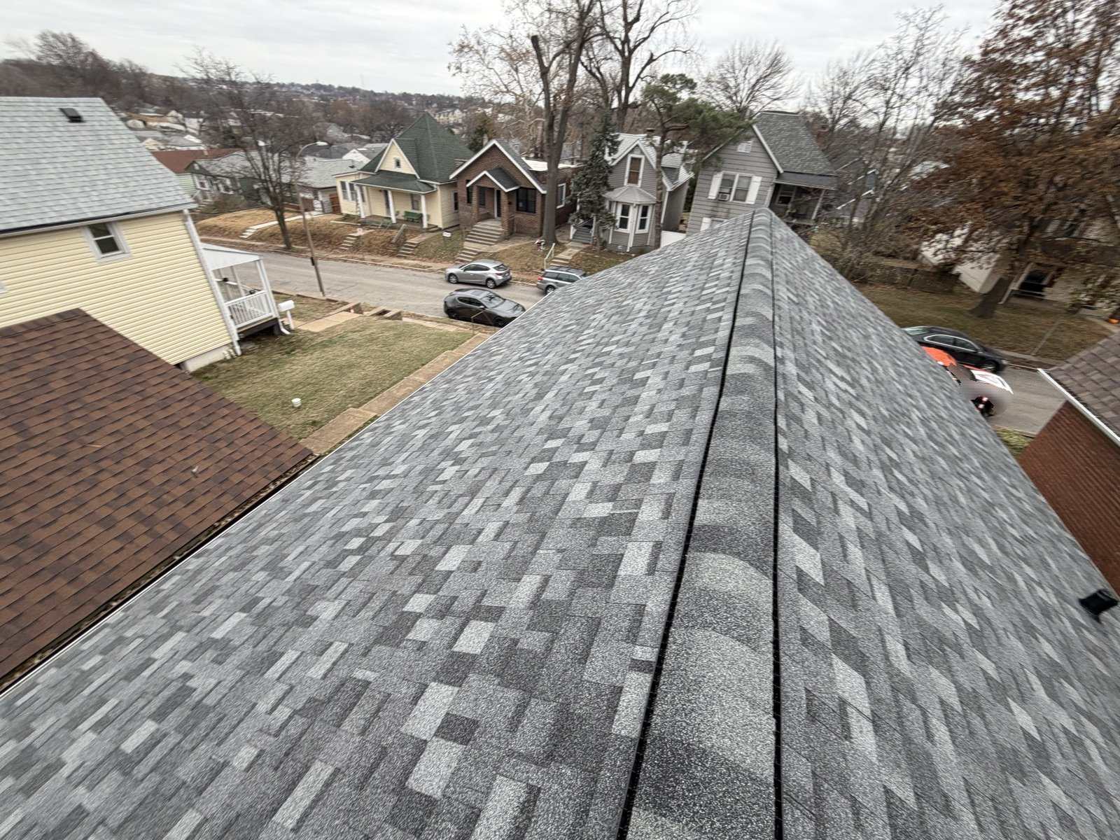 A grey architectural shingle roof with a ridge vent running away from the camera, overlooking an older St. Louis street of frame and brick houses under a winter sky
