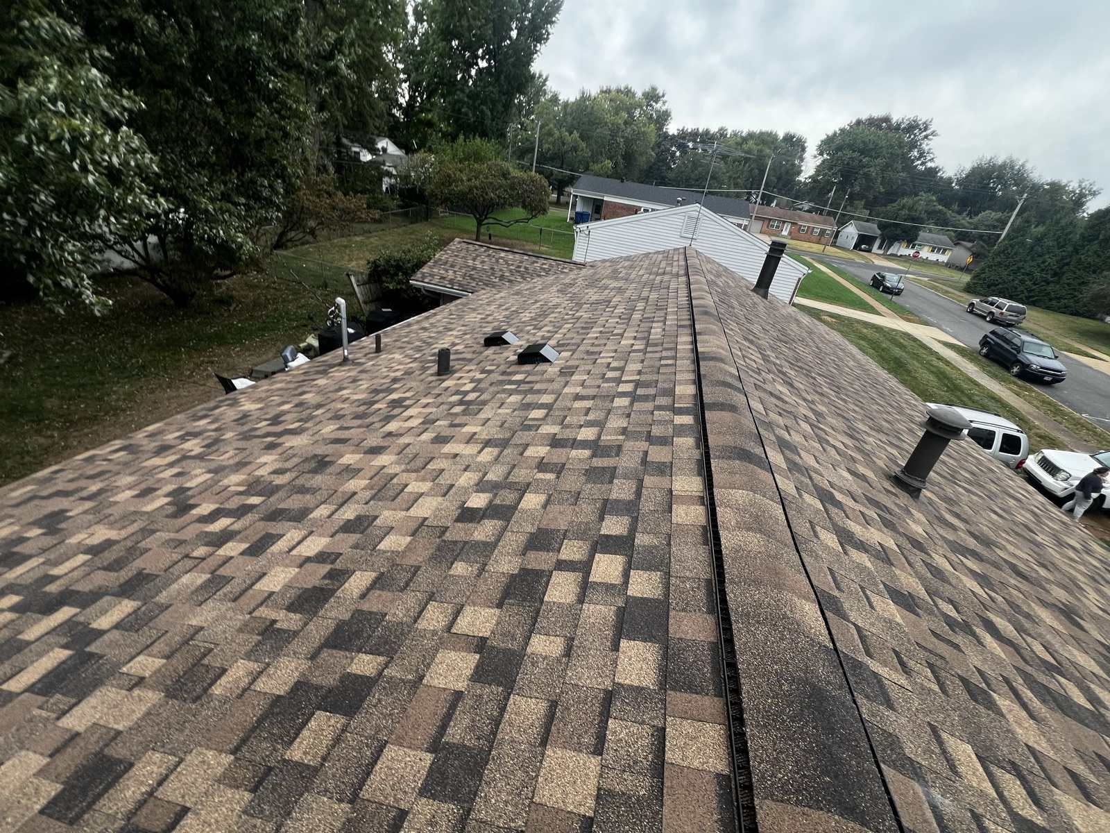 A long roof ridge with a black ridge vent, two box vents and pipe vents on the near slope, and a suburban street with parked cars below