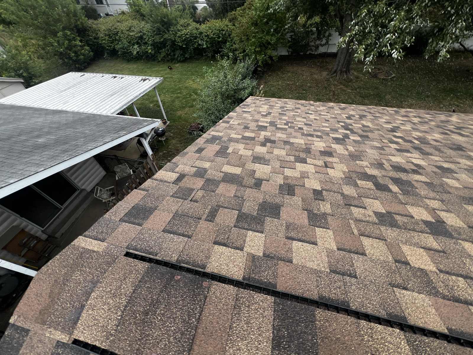 Looking down a slope of multi-tone brown architectural shingles, with a ridge vent strip along the near edge and a garden behind