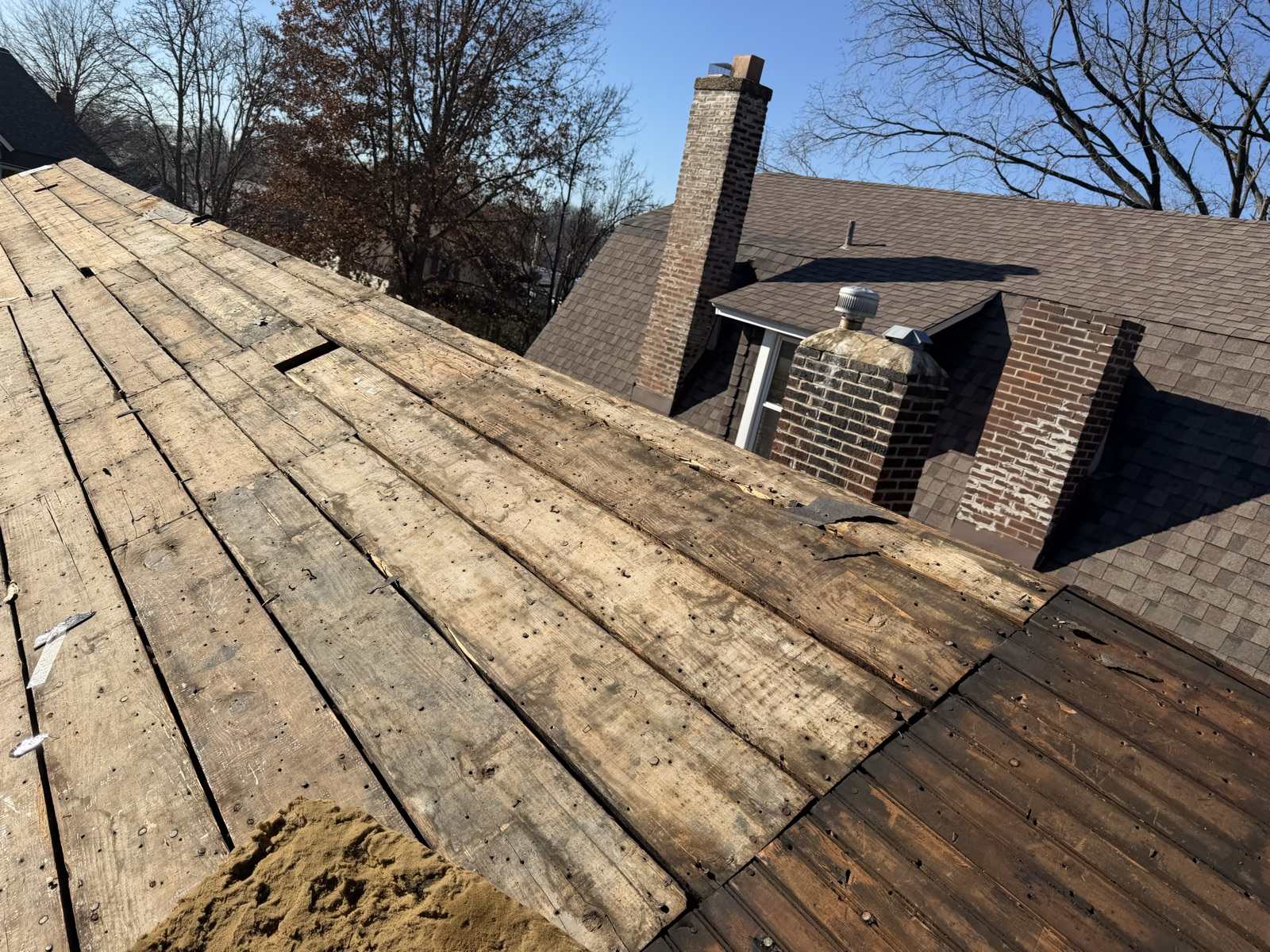 Plank decking exposed on a hip roof during a tear-off, with two brick chimneys and a turbine vent on the adjoining roof behind, and a section of noticeably darker boards at the right