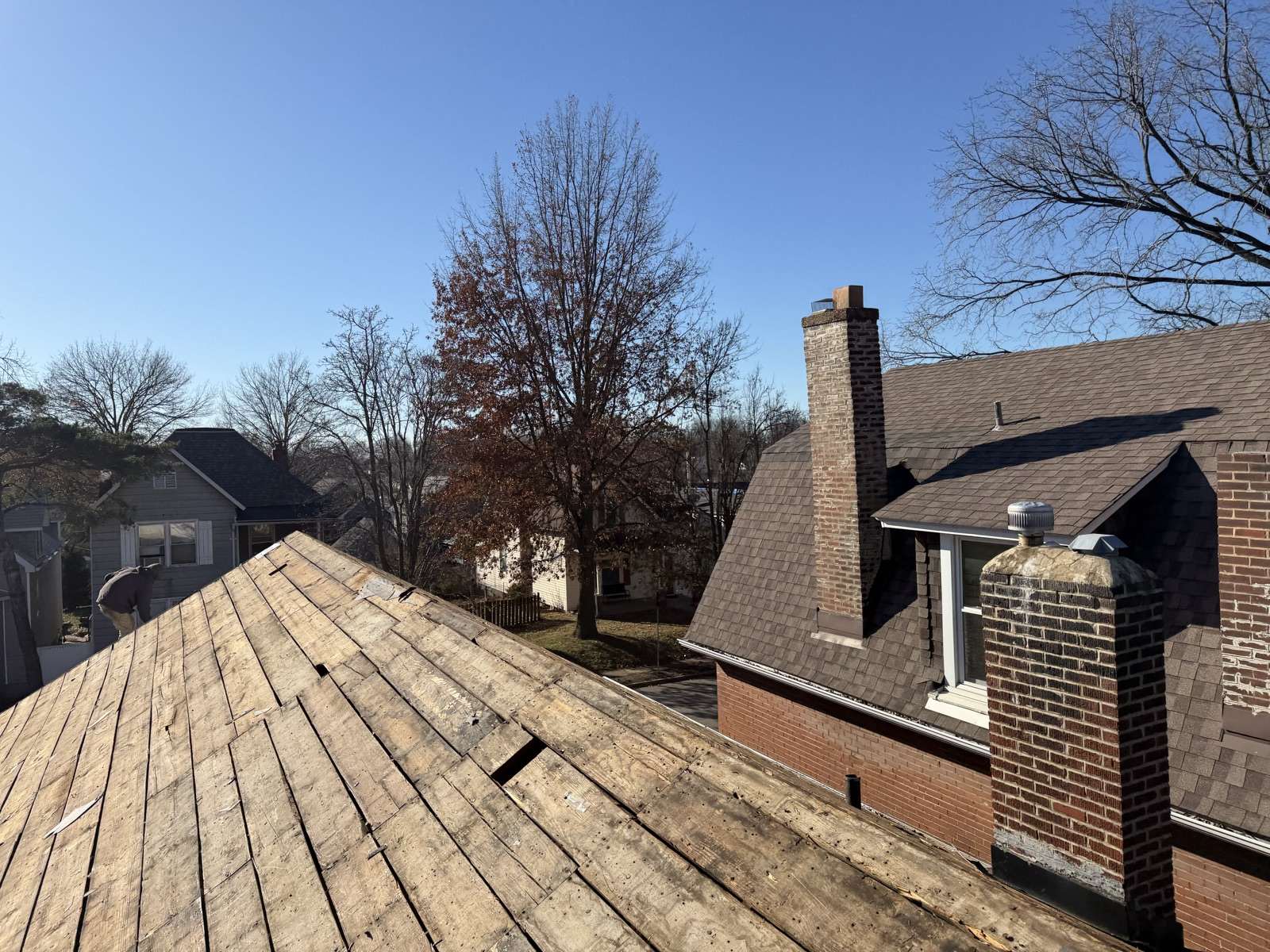 Bare wooden plank decking on a hip roof mid tear-off, with a brick chimney, a neighbouring brown shingle roof and bare winter trees behind