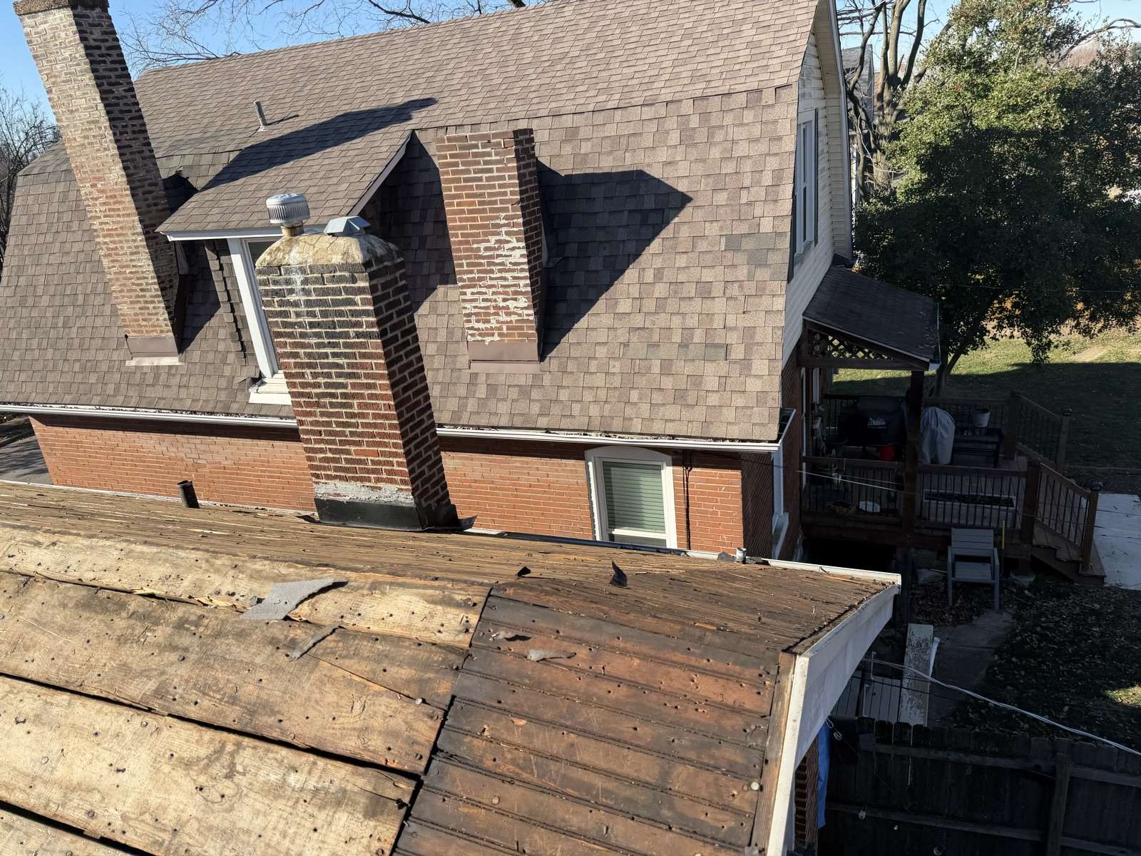 A tear-off part way through: the lower roof stripped back to plank decking while the upper slope still carries its old brown shingles, with two brick chimneys and a dormer behind